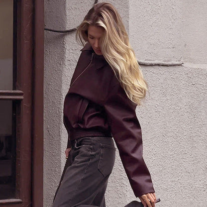 Blonde Frau in dunkelroter Lederjacke und grauer Hose lehnt an einer Wand. Modetrend, Streetstyle, Herbstmode, lässiger Look.