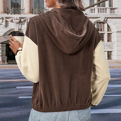 Frau in braun-beiger Kapuzenjacke mit Kaffeebecher, steht vor städtischer Kulisse. Modetrend, Herbstmode, lässiger Stil.