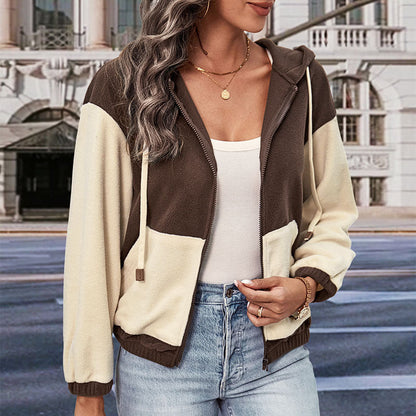 Frau in zweifarbiger Kapuzenjacke, braun und beige, mit Reißverschluss, weißem Top und Jeans, steht vor städtischer Kulisse. Mode, Streetstyle, Herbst.