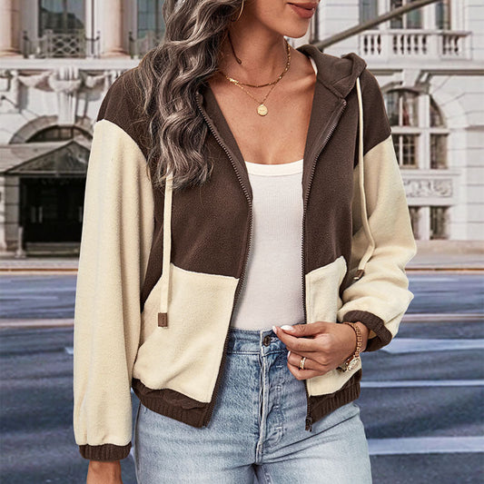 Frau in zweifarbiger Kapuzenjacke, braun und beige, mit Reißverschluss, weißem Top und Jeans, steht vor städtischer Kulisse. Mode, Streetstyle, Herbst.