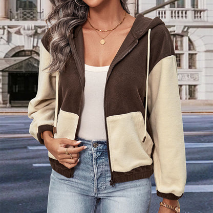 Frau in zweifarbiger Kapuzenjacke, braun und beige, mit Reißverschluss, weißem Top und blauen Jeans, steht vor städtischer Kulisse.