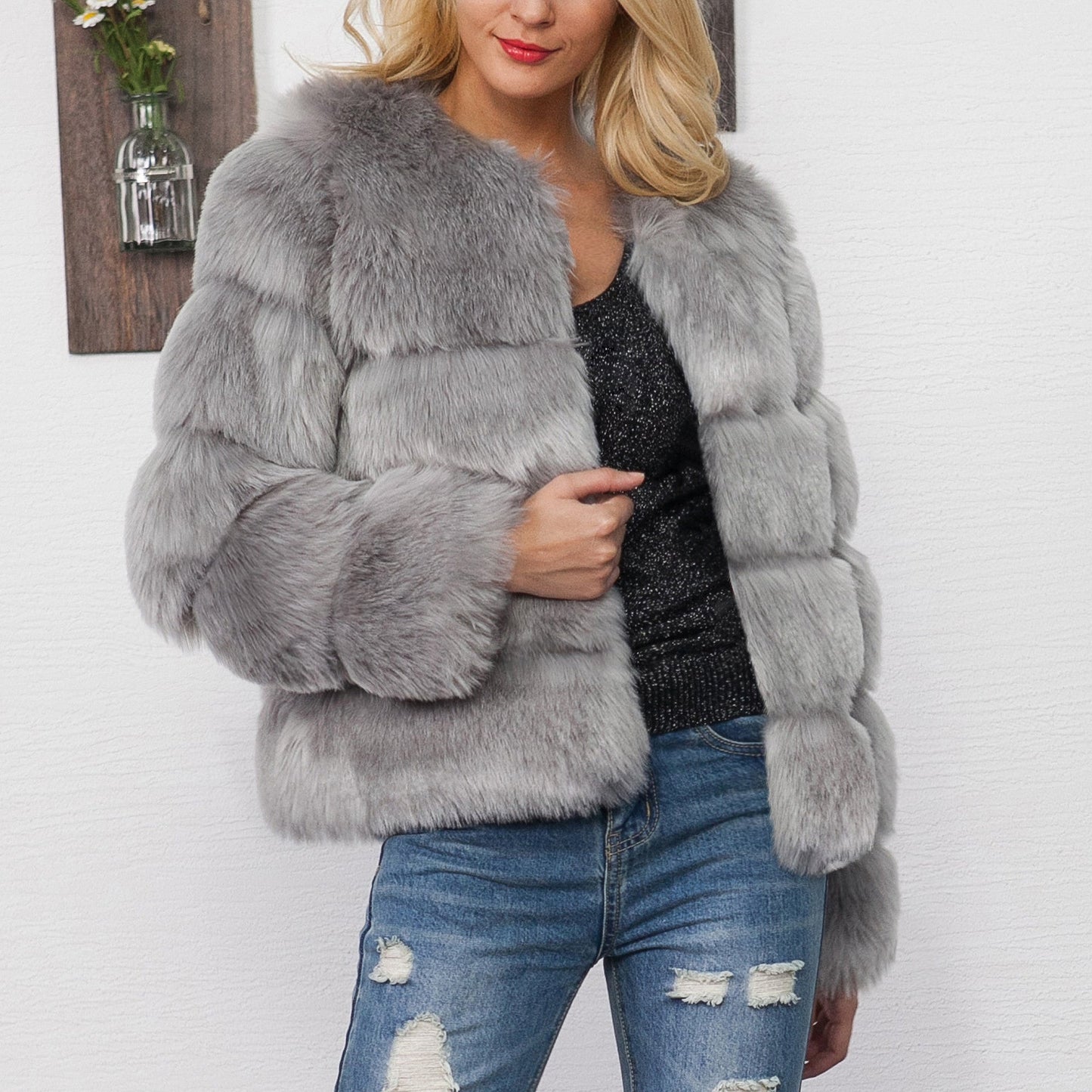 Blonde Frau in grauer Kunstpelzjacke, schwarze Glitzerbluse und zerrissenen Jeans vor weißer Wand. Modetrend, Wintermode, stilvolle Damenbekleidung.