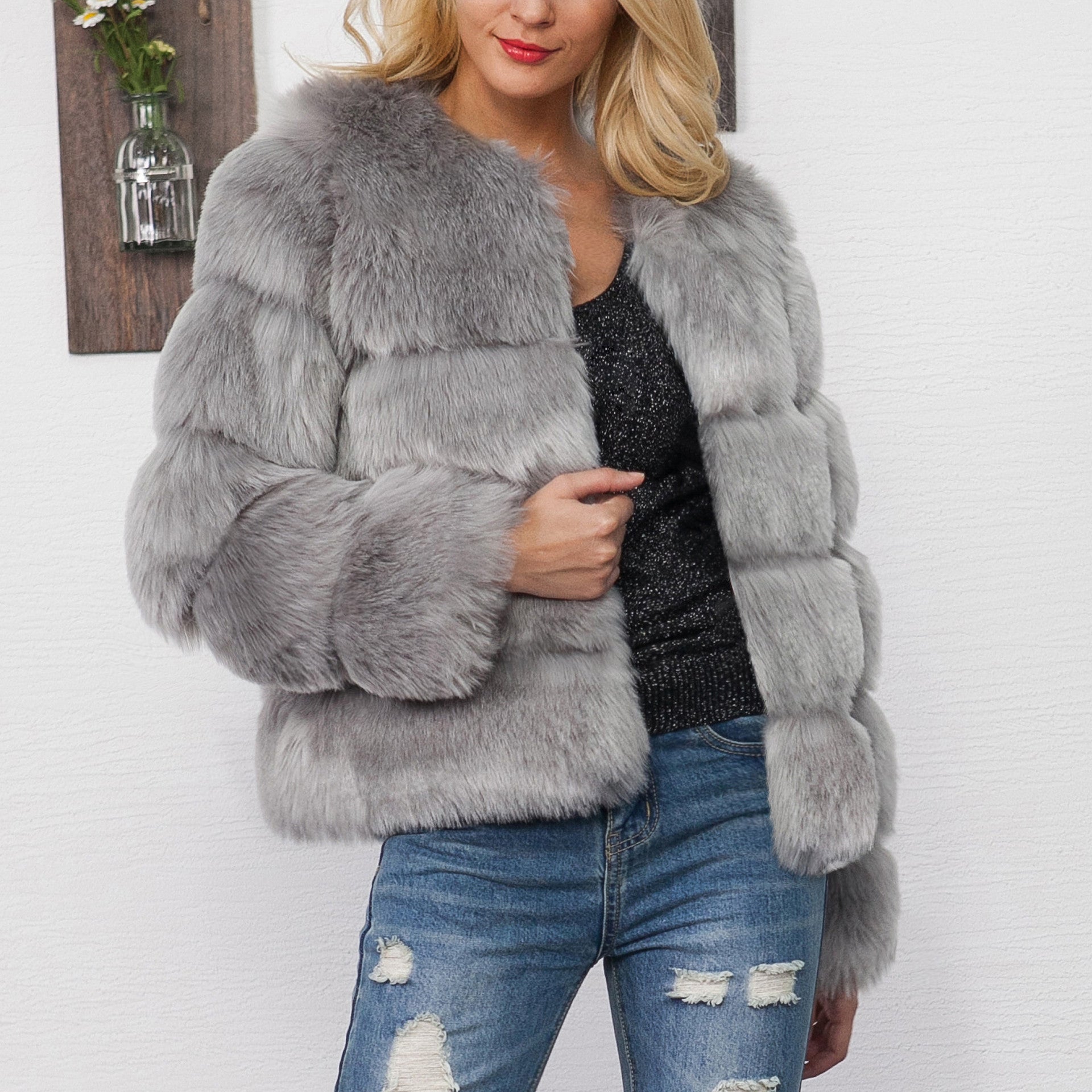 Blonde Frau in grauer Kunstpelzjacke, schwarze Glitzerbluse und zerrissenen Jeans vor weißer Wand. Modetrend, Wintermode, stilvolle Damenbekleidung.