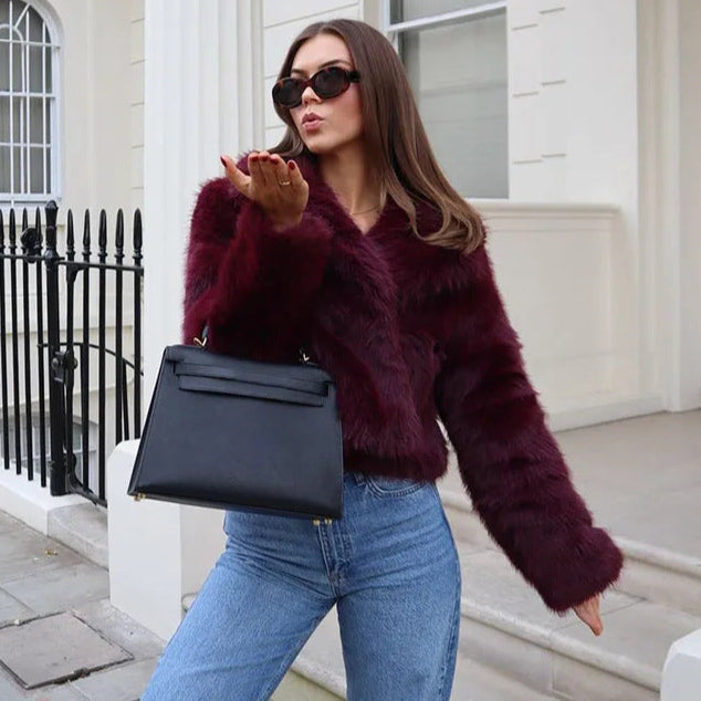 Frau in roter Kunstpelzjacke, blauer Jeans und Sonnenbrille, hält schwarze Handtasche, steht vor weißem Gebäude. Fashion, Streetstyle, Mode.
