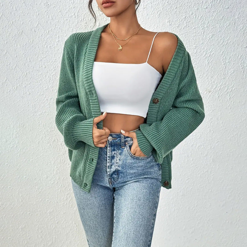 Frau in grüner Strickjacke, weißem Crop-Top und blauen Jeans vor weißer Wand. Modetrend, lässiger Stil, Damenmode, Strickjacke Outfit.