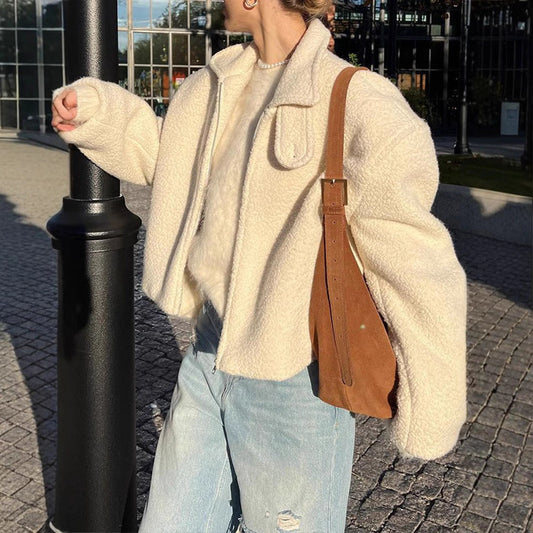 Frau in weißer Teddyjacke und blauer Jeans mit brauner Umhängetasche, steht neben einer Laterne. Herbstmode, Streetstyle, urbane Kulisse.