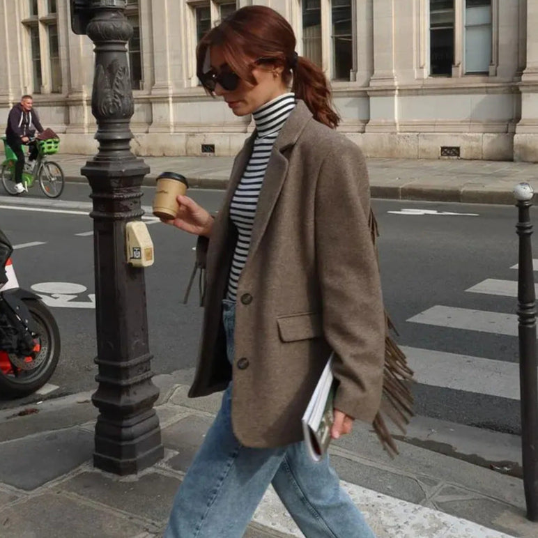 Frau im braunen Blazer und gestreiftem Rollkragenpullover überquert Straße mit Kaffeebecher und Zeitung, modischer Streetstyle, urbane Kulisse.