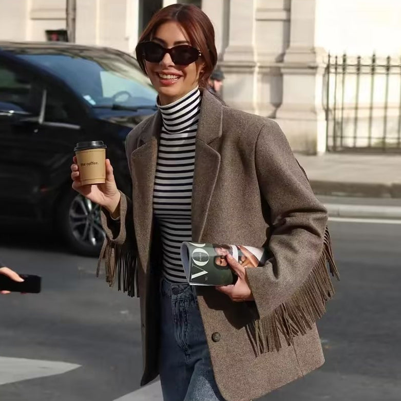 Frau in braunem Fransenmantel und Sonnenbrille hält Kaffeebecher und Zeitschrift, lächelnd auf der Straße. Modisch, urbaner Stil, Herbstmode.