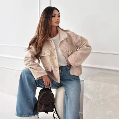 Frau in beiger Teddyjacke und blauer Schlaghose sitzt auf Hocker, hält braunen Rucksack. Modetrend, Herbstmode, Damenbekleidung, stilvoll.