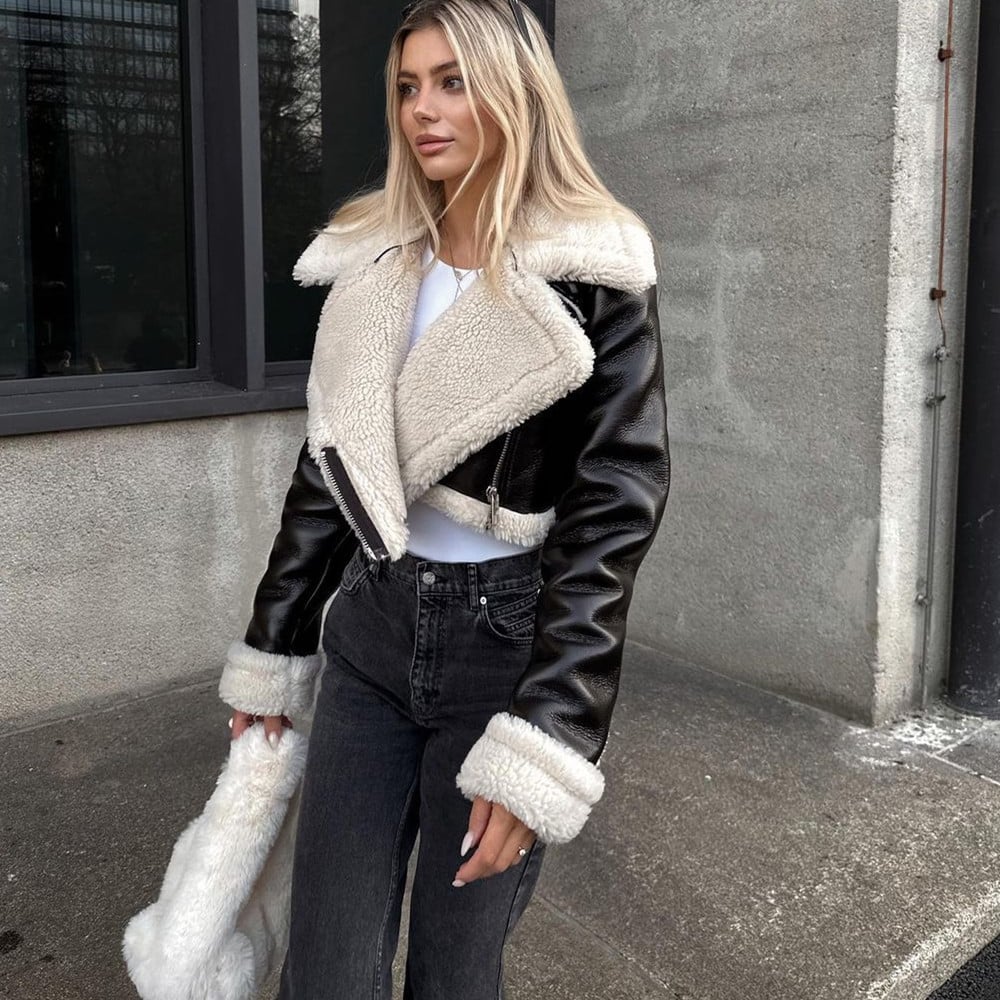 Blonde Frau in schwarzer Lederjacke mit weißem Fellkragen, weißem Top und grauer Jeans steht vor Betonwand. Wintermode, Streetstyle, trendig.