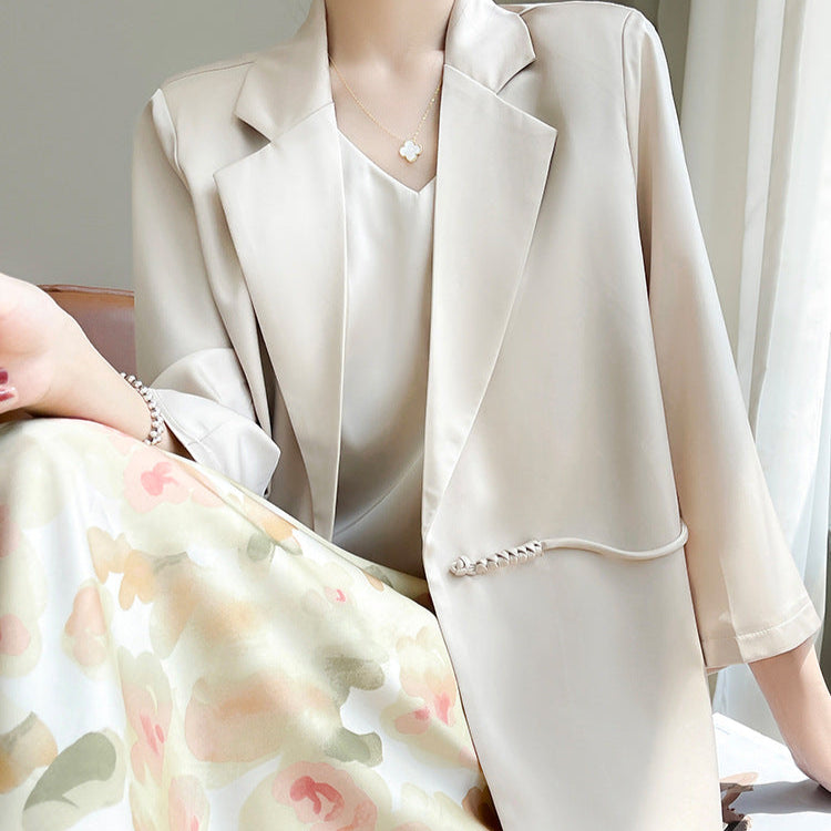 Elegante Frau in beigem Blazer und floralem Kleid, sitzt stilvoll auf einem Sessel. Mode, Damenbekleidung, schickes Outfit, Frühlingslook.