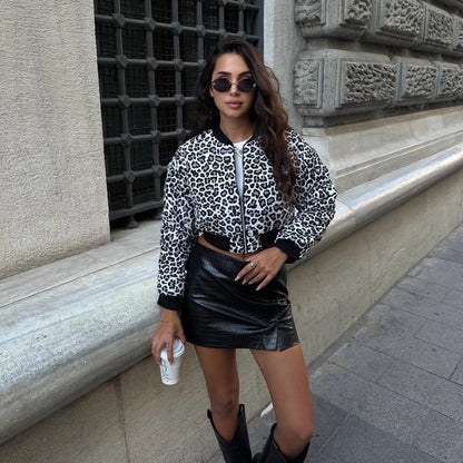 Frau in Leopardenjacke und schwarzem Lederrock, hält Kaffeebecher, steht vor historischer Steinwand, trägt Sonnenbrille und Stiefel.