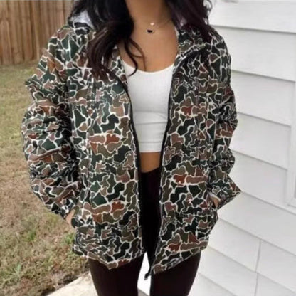 Frau in Camouflage-Jacke, weißem Top und schwarzer Hose vor weißer Wand. Modetrend, Outdoor-Bekleidung, lässiger Stil.