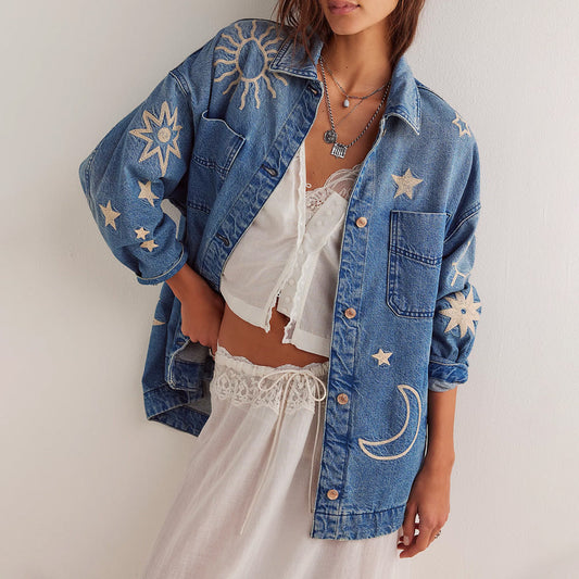 Frau trägt bestickte Jeansjacke mit Sonnen- und Mondmotiven über weißem Spitzenoberteil und Rock. Boho-Mode, lässiger Stil, trendige Damenbekleidung.