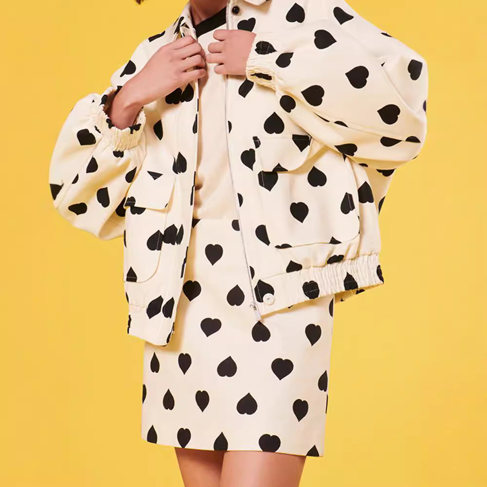 Frau in cremefarbenem Outfit mit schwarzen Herzmustern, bestehend aus Jacke und Rock, vor gelbem Hintergrund. Modetrend, Herzmuster, Damenmode.