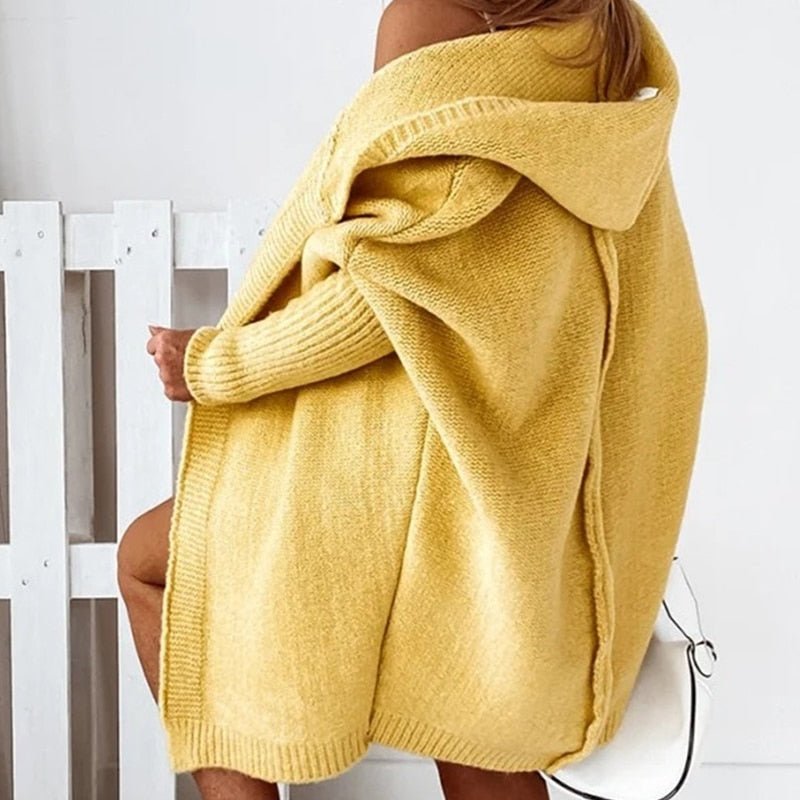 Frau in gelbem Strickmantel mit Kapuze, sitzt auf weißem Stuhl. Modetrend Herbst, gemütlicher Oversize-Look, stilvolle Damenbekleidung.