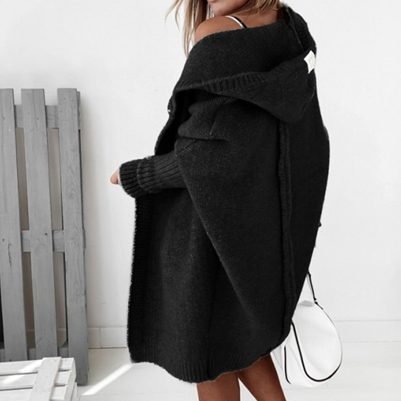 Frau in schwarzem Strickmantel mit Kapuze, seitlich stehend, hält weiße Tasche. Modetrend Herbst/Winter, gemütlicher Oversize-Look.