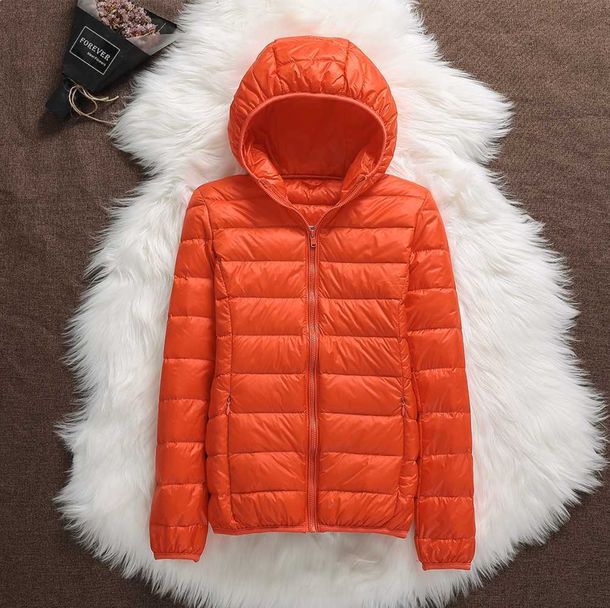Orangefarbene Daunenjacke mit Kapuze auf weißem Fellteppich, modische Winterbekleidung, warm und leicht, ideal für kalte Temperaturen.