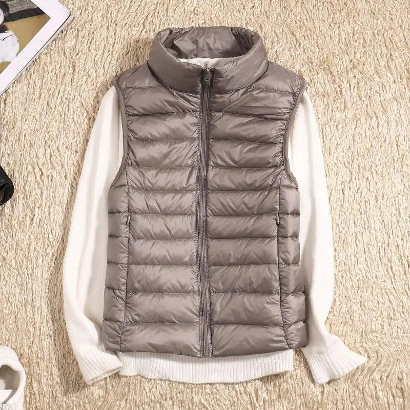 Graue Daunenweste mit Reißverschluss auf cremefarbenem Pullover, flach auf beigem Teppich liegend. Modetrend, Herbstbekleidung, Damenmode.
