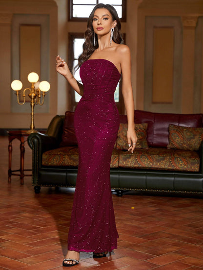 Frau in elegantem, funkelndem, bordeauxrotem Abendkleid, steht in stilvollem Wohnzimmer mit Ledersofa und Stehlampe. Elegante Abendmode, Glamour.