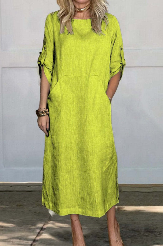 Frau in knielangem, grünem Leinenkleid mit Taschen und hochgekrempelten Ärmeln, steht vor weißer Wand. Modetrend, Sommerkleid, Damenmode.