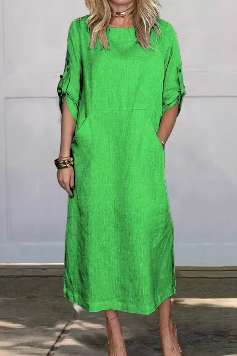 Frau in grünem Leinenkleid, knielang, mit hochgekrempelten Ärmeln, steht vor grauer Wand. Modetrend, Sommerkleid, Damenmode.