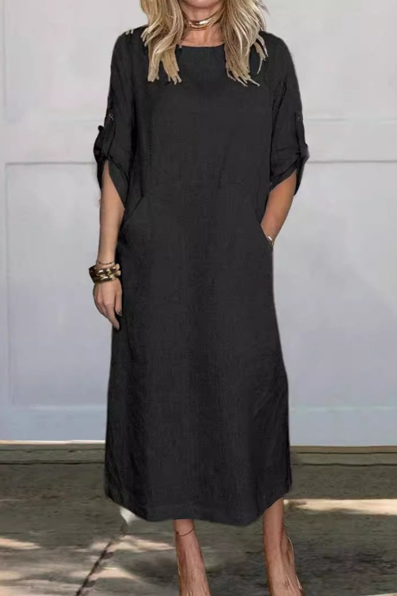 Frau in schwarzem, knielangem Kleid mit halblangen Ärmeln und Taschen, steht vor grauer Wand. Modisches Damenoutfit, lässiger Stil.