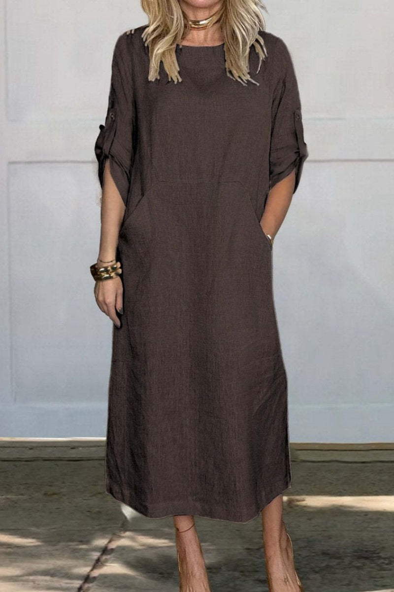 Frau in braunem Leinenkleid mit Taschen, knielang, lässiger Stil, steht vor weißer Wand. Perfekt für Sommermode und Freizeitkleidung.