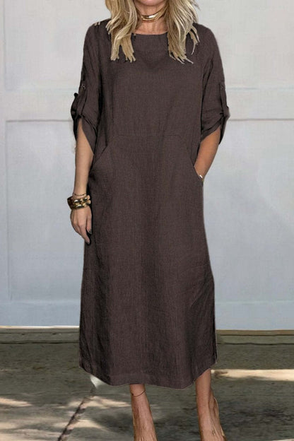 Frau in braunem Leinenkleid mit Taschen, knielang, lässiger Stil, steht vor weißer Wand. Perfekt für Sommermode und Freizeitkleidung.