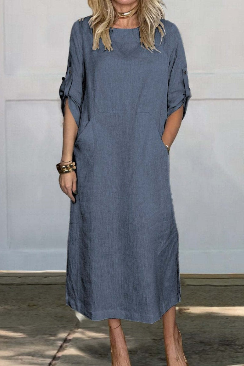 Frau in lockerem, knielangem, blauem Leinenkleid mit hochgekrempelten Ärmeln und Taschen, steht vor weißer Wand. Modetrend, Sommerkleid, Damenmode.