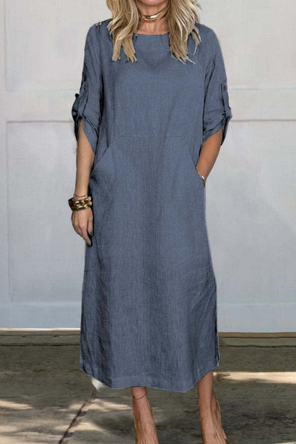 Frau in lockerem, knielangem, blauem Leinenkleid mit hochgekrempelten Ärmeln und Taschen, steht vor weißer Wand. Modetrend, Sommerkleid, Damenmode.
