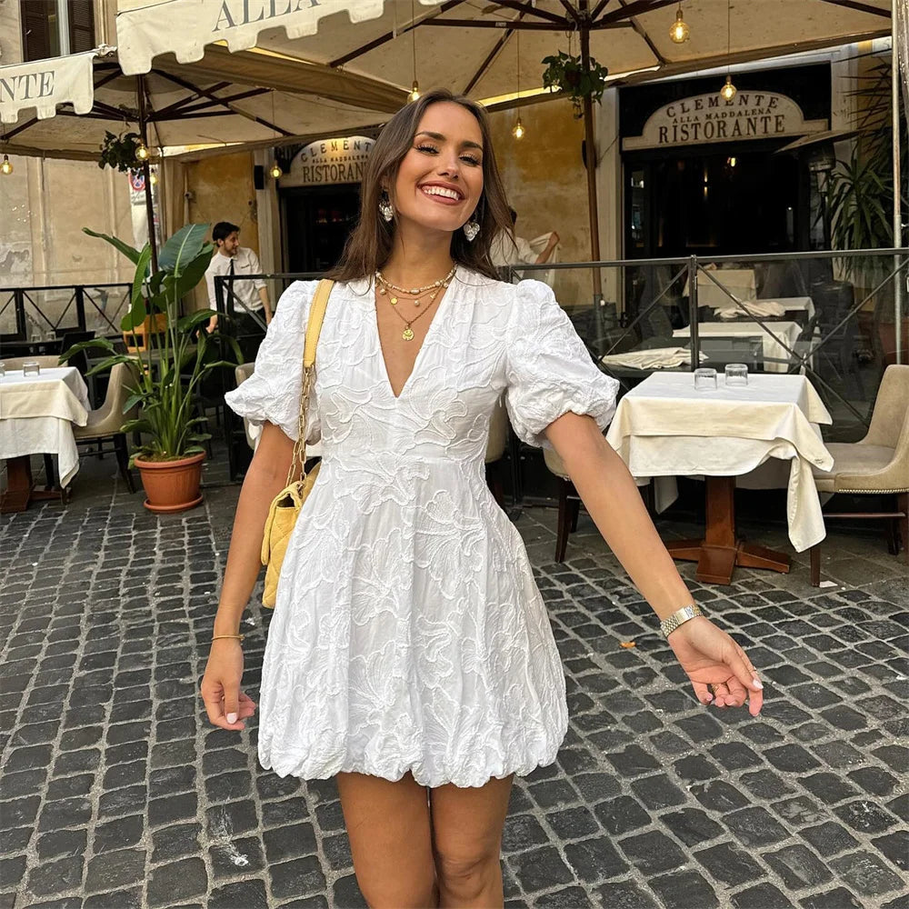 Eine lächelnde Frau in einem weißen Sommerkleid steht auf einer gepflasterten Straße vor einem italienischen Restaurant mit Außenterrasse.