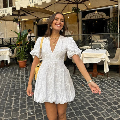 Eine lächelnde Frau in einem weißen Sommerkleid steht auf einer gepflasterten Straße vor einem italienischen Restaurant mit Außenterrasse.