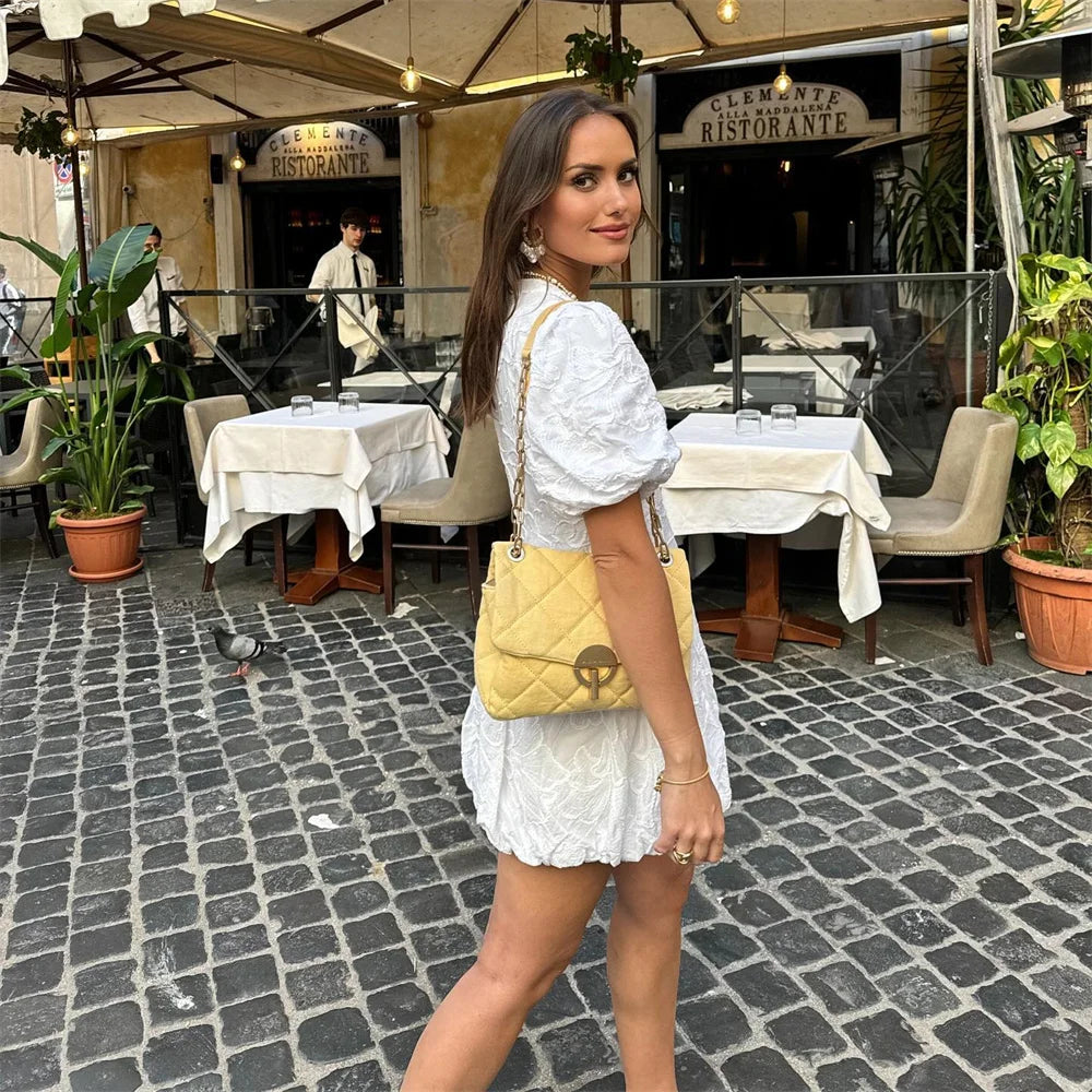 Eine Frau in einem weißen Kleid steht auf Kopfsteinpflaster vor einem Restaurant. Sie trägt eine gelbe Designer-Handtasche.