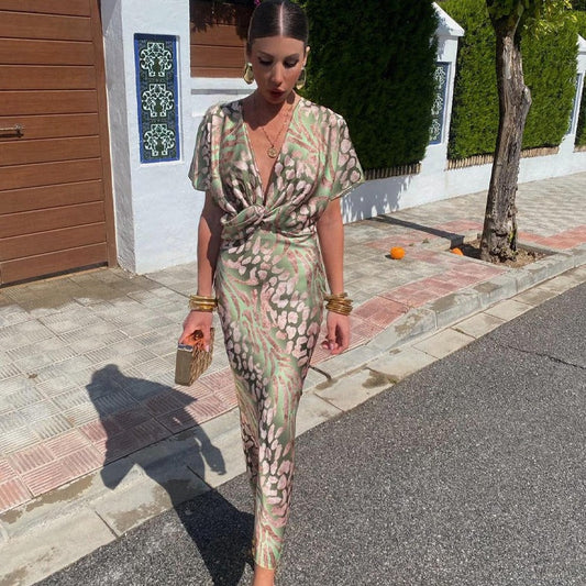 Frau in elegantem, gemustertem Kleid spaziert auf Bürgersteig, trägt goldene Accessoires und Clutch, sonniger Tag, stilvolle Mode.