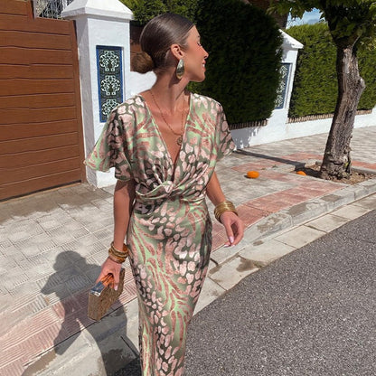 Frau in elegantem, gemustertem Kleid auf Gehweg, trägt goldene Accessoires und hält eine kleine Tasche. Sommermode, stilvoll, schick.