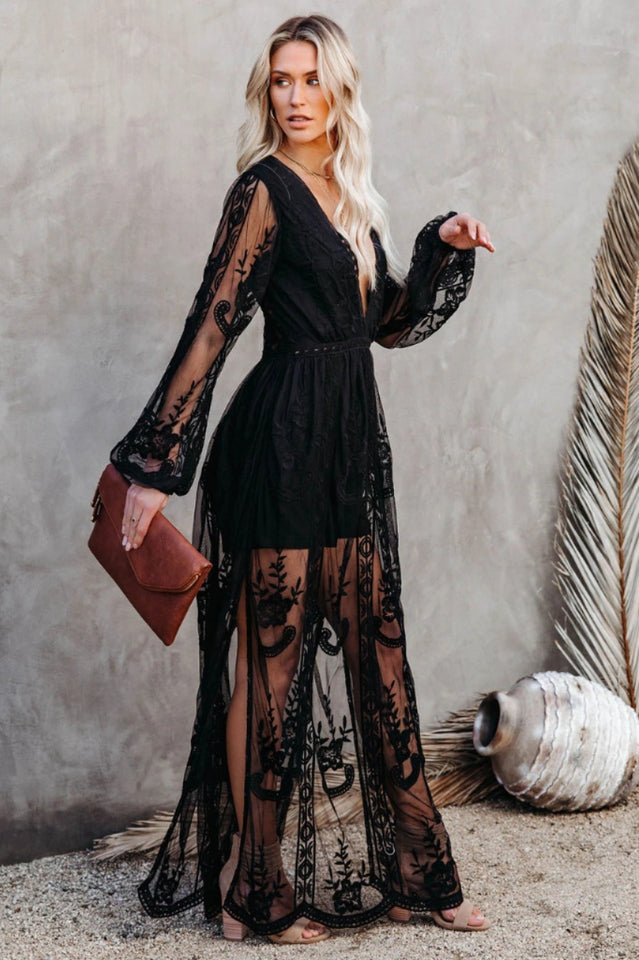 Blonde Frau in schwarzem, transparentem Spitzenkleid mit langen Ärmeln, hält braune Clutch, steht vor neutralem Hintergrund mit Deko-Elementen.