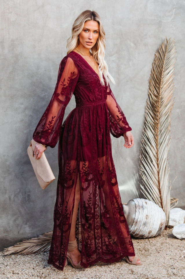 Frau in elegantem, langem, bordeauxrotem Spitzenkleid mit transparenten Ärmeln, steht vor grauer Wand, hält eine beige Clutch.