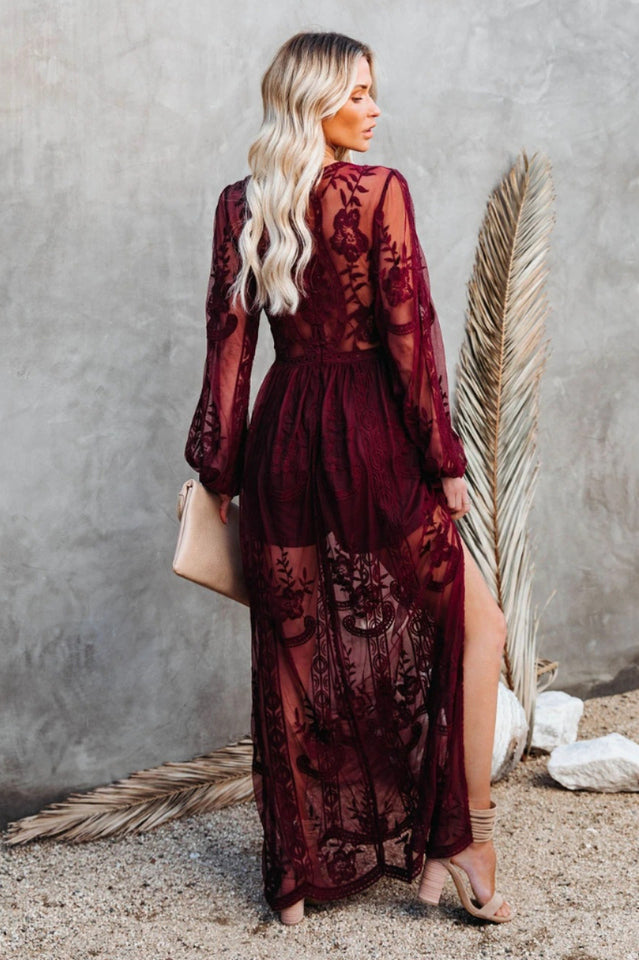 Frau in langem, transparentem, bordeauxrotem Spitzenkleid mit floralen Mustern, steht vor grauer Wand, hält beigefarbene Clutch, trägt Sandaletten.
