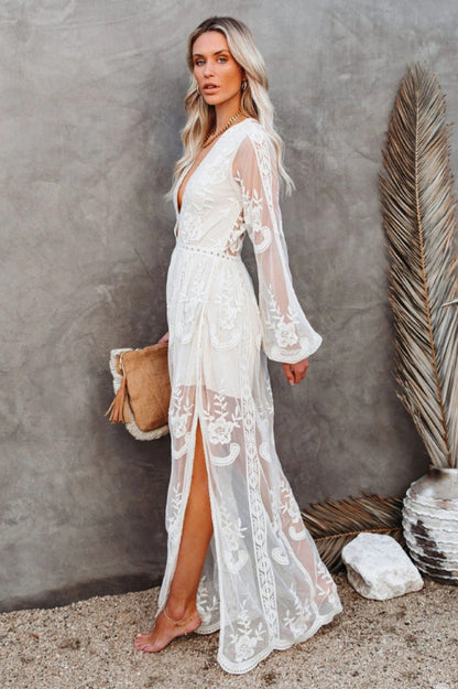 Frau in weißem Spitzenkleid, barfuß auf Sand, mit brauner Clutch. Elegantes Sommeroutfit, Boho-Stil, ideal für Strandhochzeiten oder Festivals.