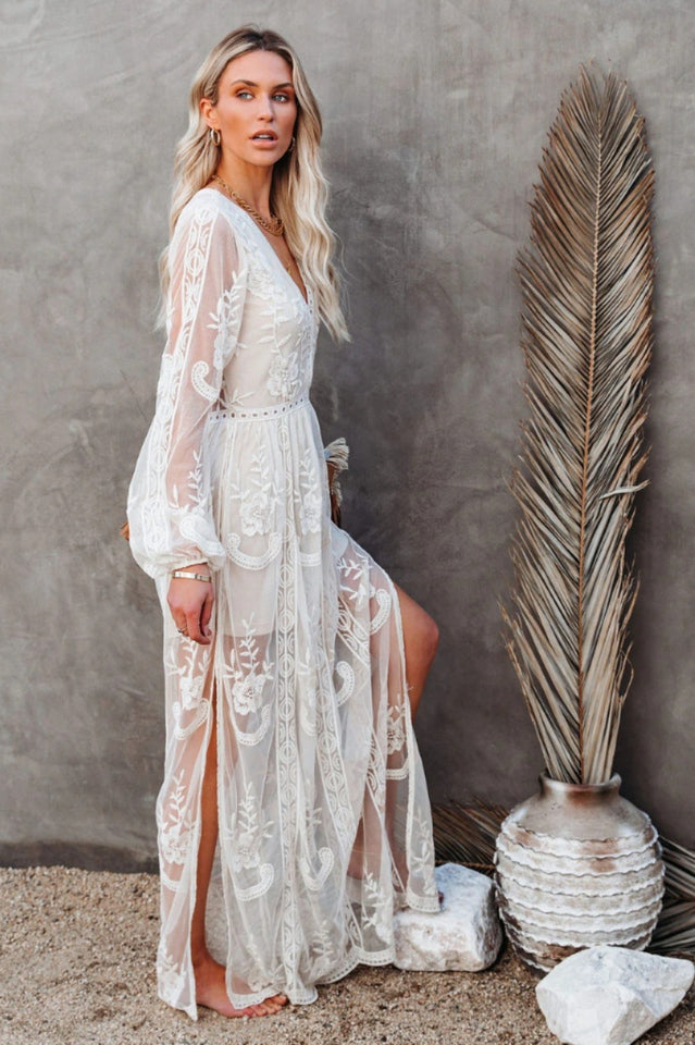 Frau in weißem Spitzenkleid posiert neben großer Trockenpflanze und Keramikvase vor grauer Wand. Elegante Mode, Boho-Stil, Sommerkleid.