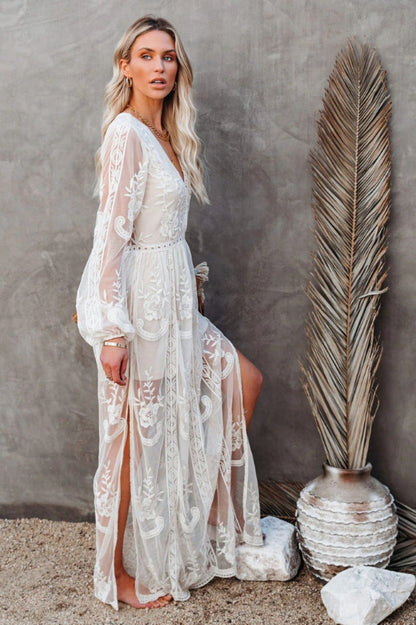 Frau in weißem Spitzenkleid posiert neben großer Trockenpflanze und Keramikvase vor grauer Wand. Elegante Mode, Boho-Stil, Sommerkleid.
