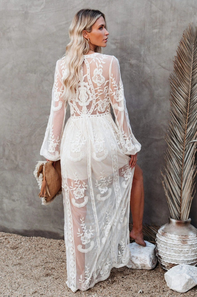 Frau in weißem, transparentem Spitzenkleid vor grauer Wand, mit beigem Handtasche. Elegante Mode, Sommerkleid, Boho-Stil, Damenmode.