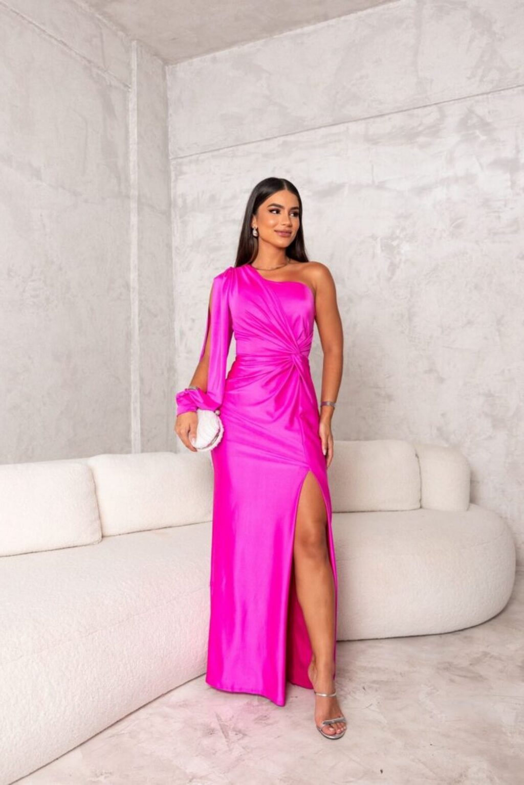 Frau in einem leuchtend pinken, schulterfreien Abendkleid mit hohem Beinschlitz, steht vor einer modernen, minimalistischen Betonwand.