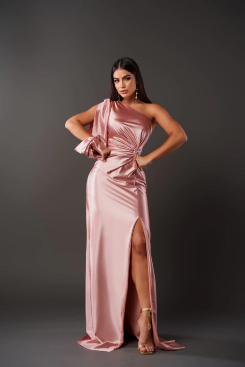 Frau in elegantem, rosafarbenem Abendkleid mit asymmetrischem Schnitt und hohem Beinschlitz, posiert vor grauem Hintergrund.