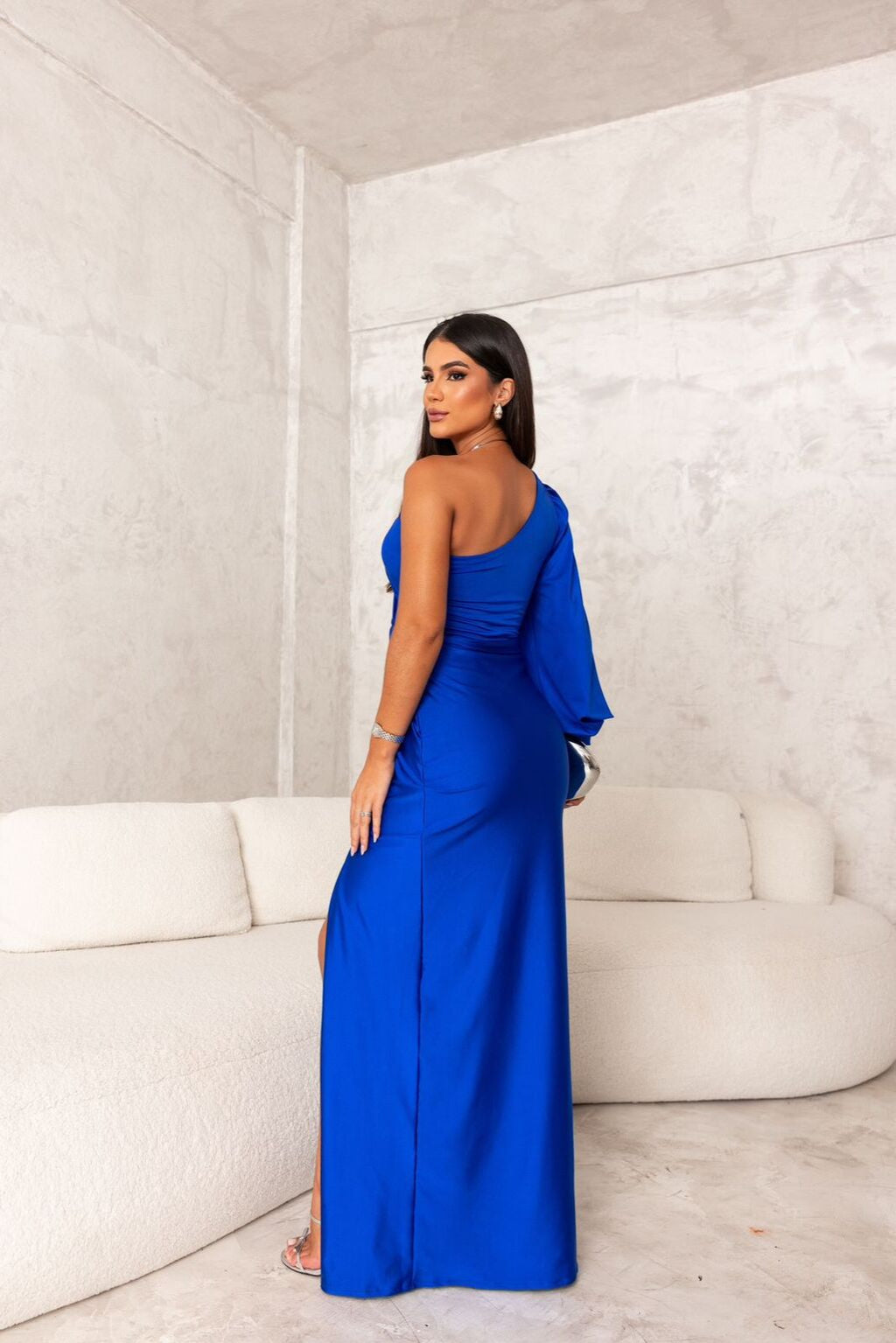 Frau in elegantem, blauem Abendkleid mit asymmetrischem Schnitt, posiert in modernem, minimalistischem Raum mit weißem Sofa.