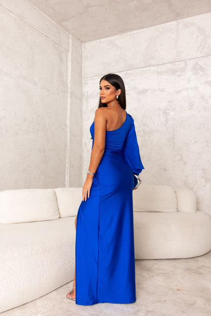 Frau in elegantem, blauem Abendkleid mit asymmetrischem Schnitt, posiert in modernem, minimalistischem Raum mit weißem Sofa.