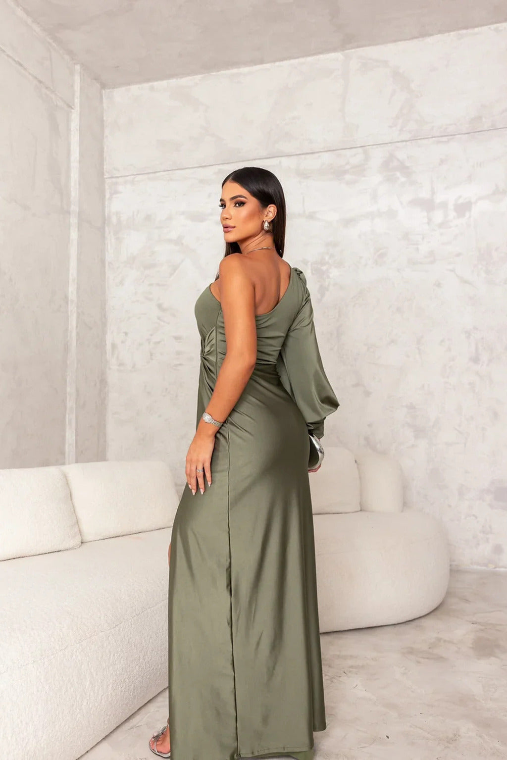Frau in elegantem, olivgrünem Abendkleid mit asymmetrischem Design steht in modernem, minimalistischem Raum mit weißem Sofa.