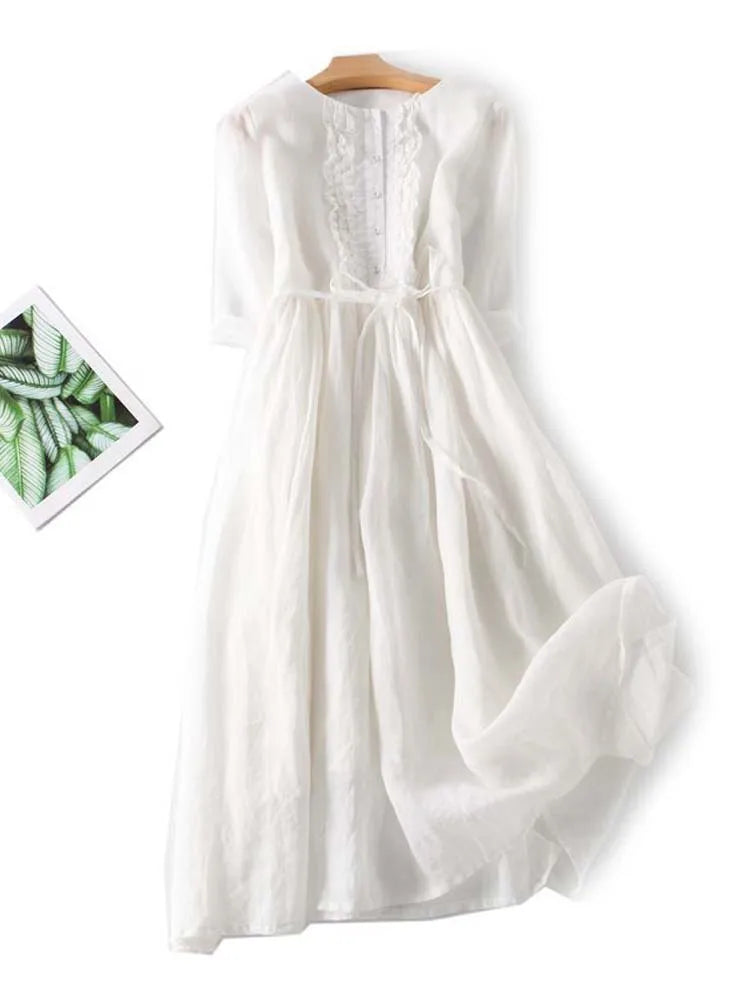 Weißes, langes Sommerkleid mit Spitzenbesatz und Bindegürtel auf Holzkleiderbügel, daneben ein Bild mit grünen Blättern. Ideal für elegante Anlässe.