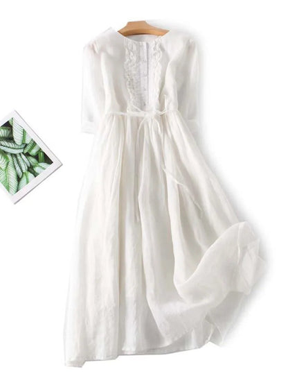 Weißes, langes Sommerkleid mit Spitzenbesatz und Bindegürtel auf Holzkleiderbügel, daneben ein Bild mit grünen Blättern. Ideal für elegante Anlässe.